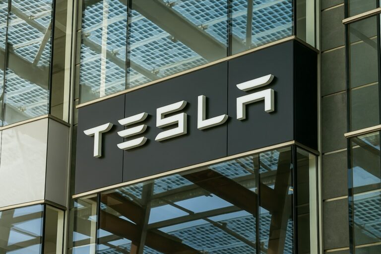 Tesla building facade featuring a bold, modern Tesla logo in white 3D lettering on a sleek black signboard, with large glass panels and a solar-panel-covered roof reflecting the sky.